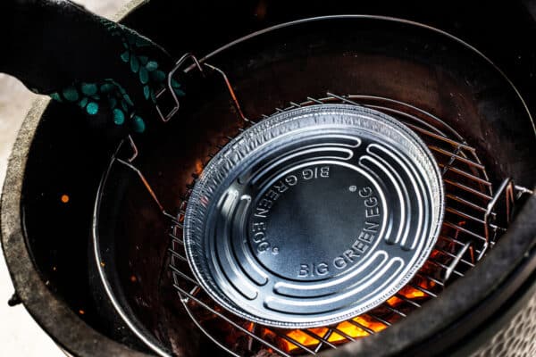 Big Green Egg grill belső nézete, hőálló kesztyűvel elhelyezett alumínium csepegtető tálca izzó parázs fölé, rozsdamentes rácson