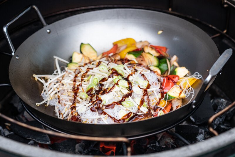 Big Green Egg szénacél wokban pirított zöldségek grillezése kültéren, fa spatulával keverve.