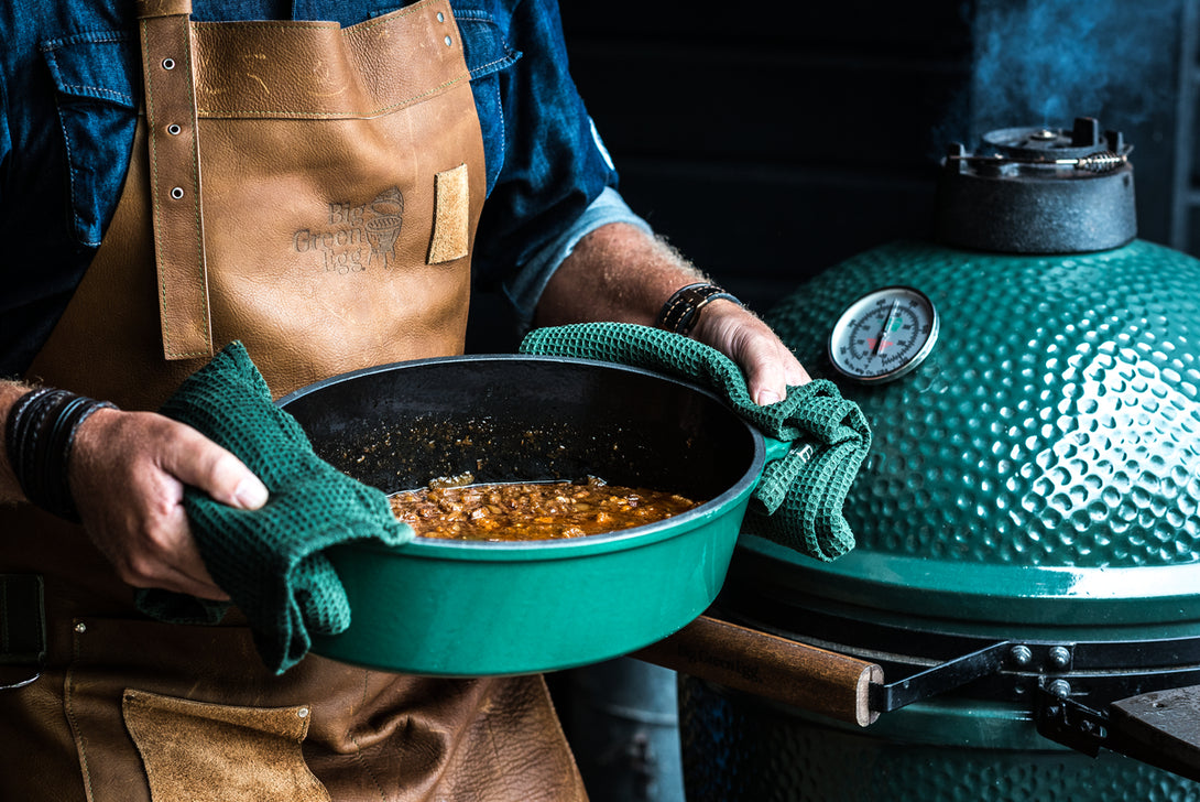 Big Green Egg ovális öntöttvas lábasban készülő étel kültéri grillezés során, zöld kerámia grill mellett.