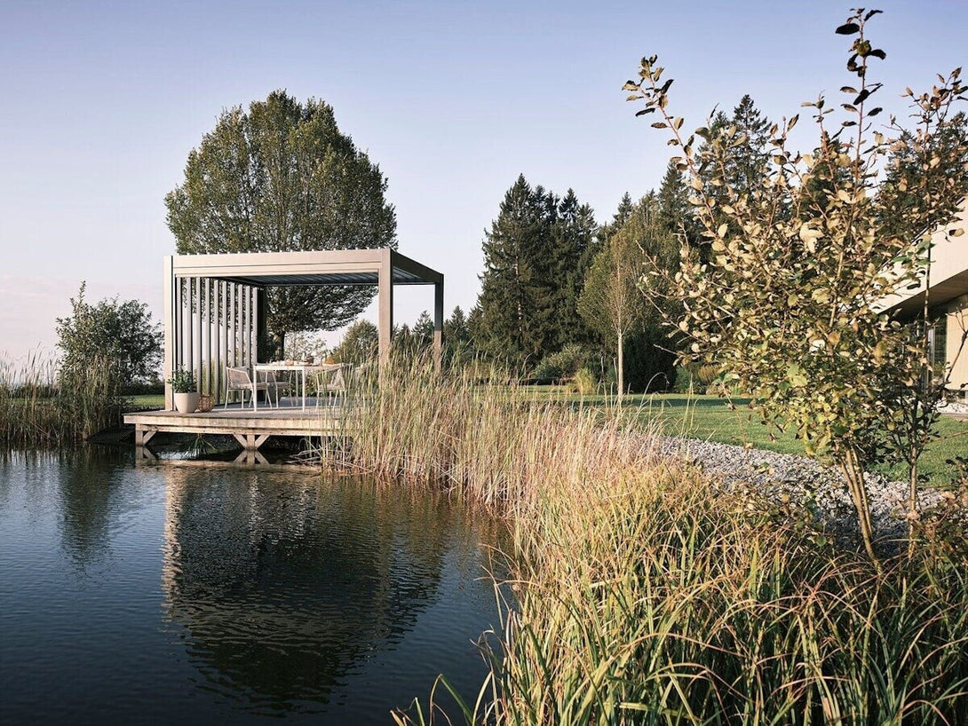 Biohort pergola ezüstmetál színben, tóparti stégen elhelyezve, modern kerti étkezőbútorokkal berendezve, természetközeli környezetben.
