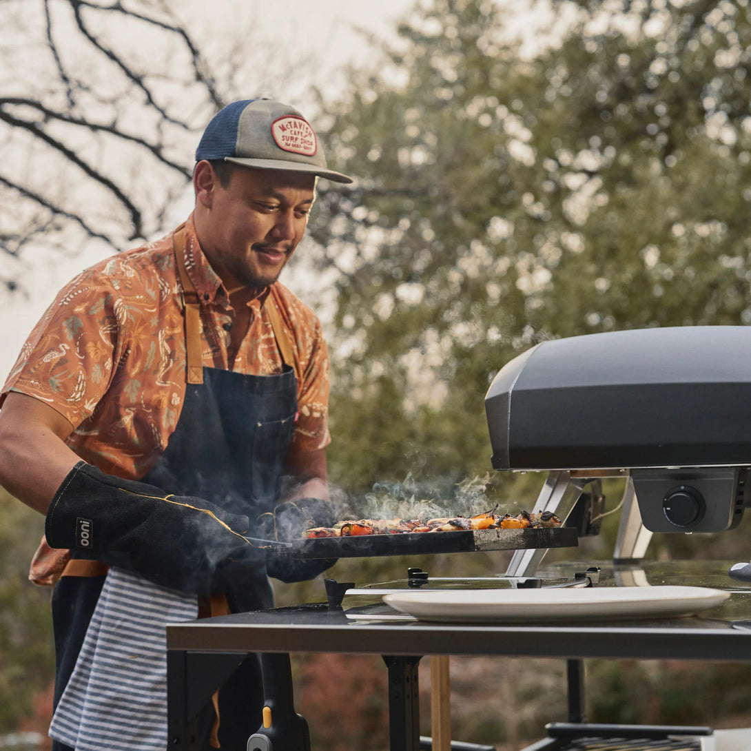 Grillételek sütése Ooni Koda 2 Pro kemencében egy kerti asztalon, séffel a háttérben

