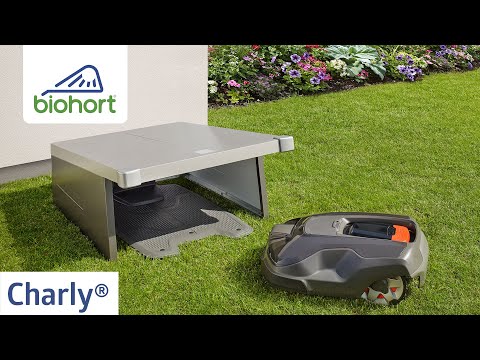 Biohort Charly robotfűnyíró garázs promóciós videóból – modern, esztétikus fémkonstrukció zöld gyepen, mellette robotfűnyíró, háttérben virágágyás és a Biohort logó.